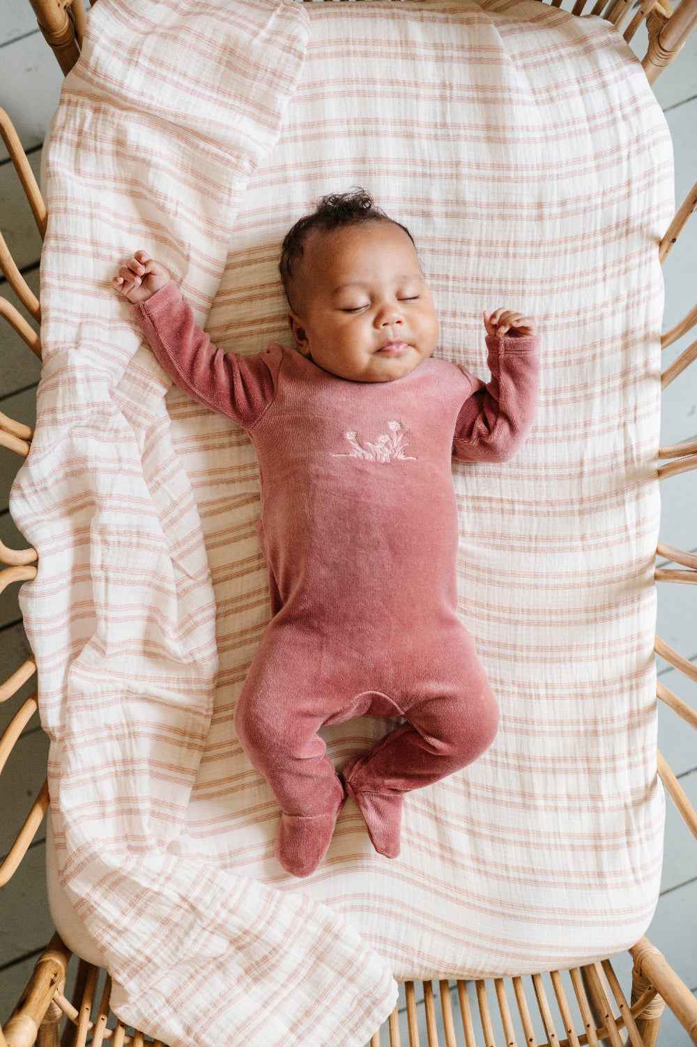 A sleeping newborn baby wearing a dusty rose pink footed sleeper, lying on their back on a cream and pink striped muslin blanket inside a wicker basket.