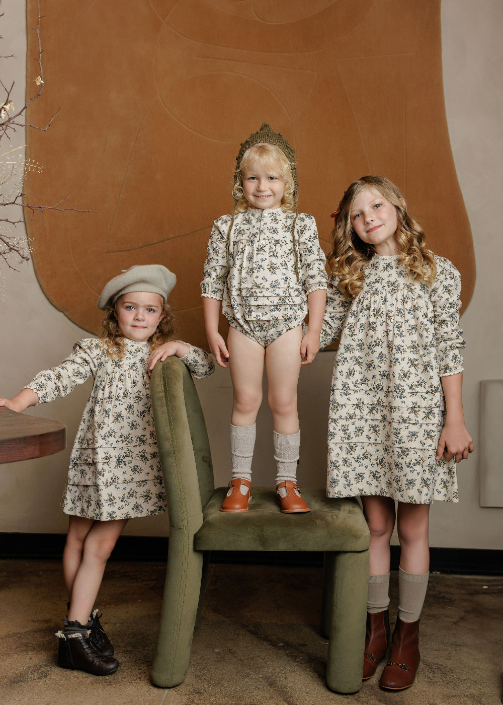 Three young girls in matching floral outfits standing in a room with a brown wall and green chair.